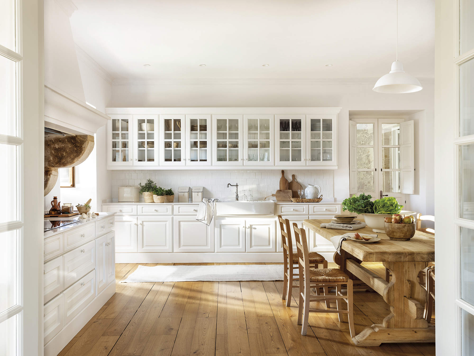 cocina con puertas acrisoladas blancas, suelo de madera natural con muebles blancos, mesa y sillas de madera, plantas aromáticas